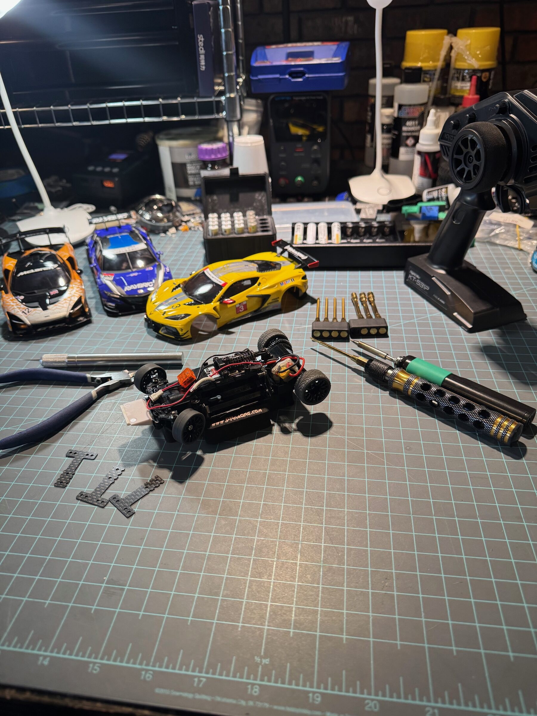 Mini-Z pit setup with stripped MR-03 chassis, three body shells, tools, and transmitter on the bench